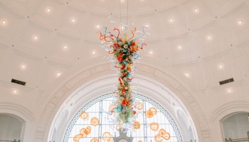 Colorful glass art chandelier hanging from a high, ornate ceiling in a bright, elegant public space or gallery.
