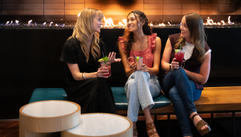Three women are sitting and chatting by a fireplace, enjoying colorful drinks and smiling.