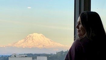 A woman looks out a window at a snow-capped mountain, likely Mount Rainier, during sunrise or sunset in this serene scene.