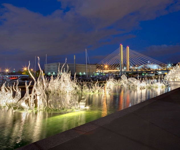 The image shows an illuminated glass sculpture installation by a waterfront at night, with a bridge and city lights in the background.