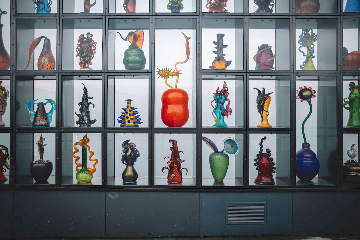 The image shows a display case filled with various colorful, artistic glass sculptures in different shapes and designs, organized in individual cubicles.