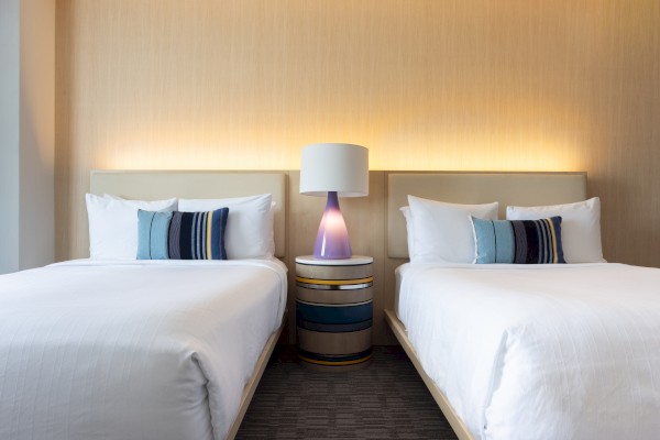 Two neatly made hotel beds with striped pillows, separated by a colorful bedside table and lamp, against a warm wooden wall background.