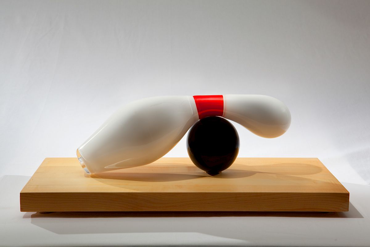 The image shows a sculptural arrangement of two bent bowling pins balancing on a black ball, all placed on a wooden base.