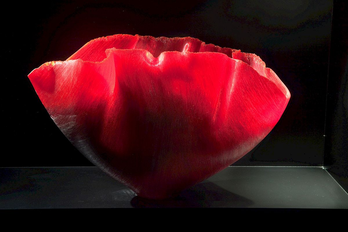 The image shows a large, vibrant red bowl with a jagged rim, likely made of glass or some other translucent material, against a dark background.