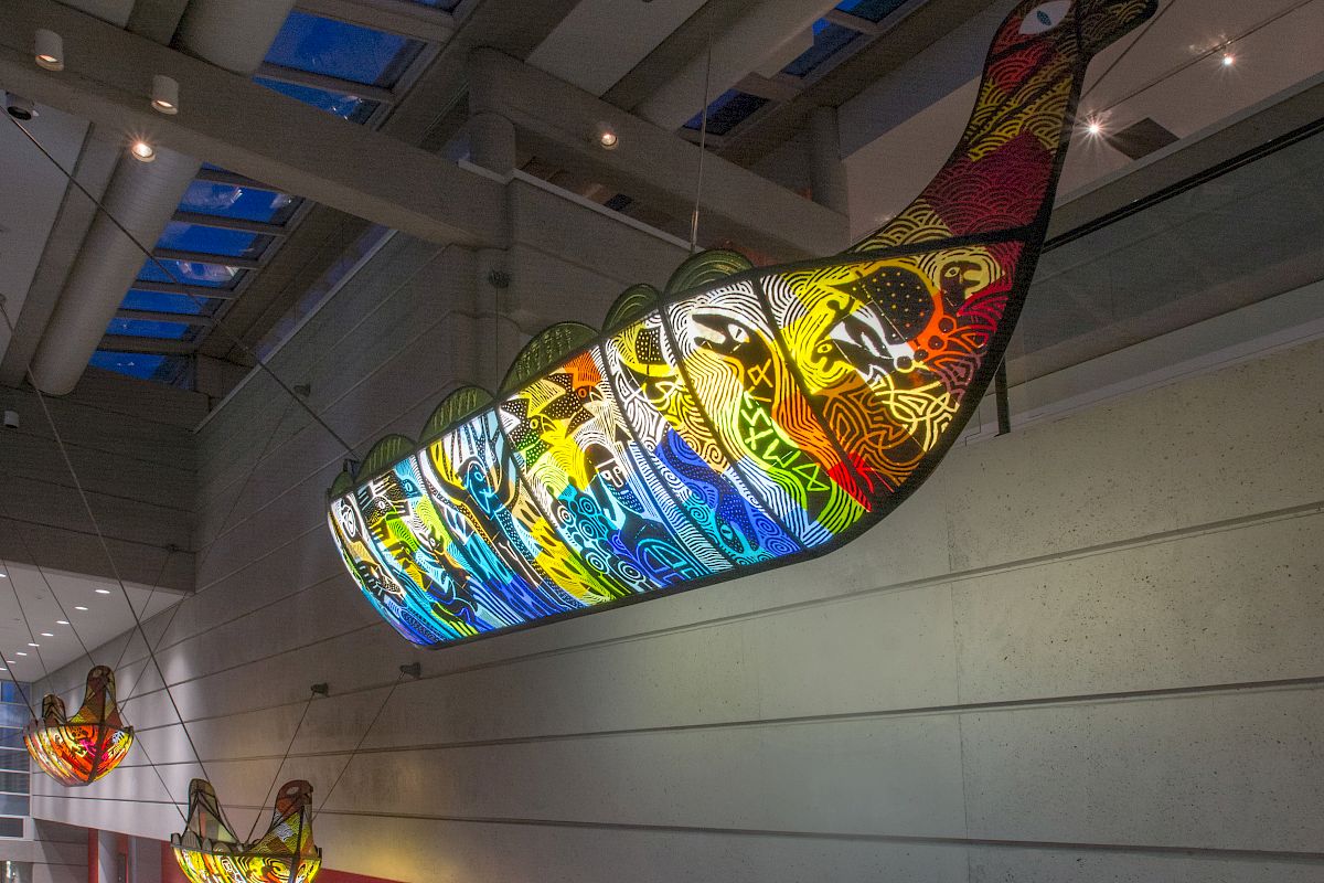 The image shows colorful, bird-shaped light fixtures hanging from the ceiling in an indoor space with skylights and contemporary architectural design.