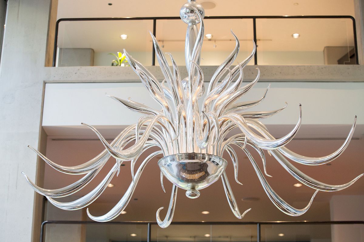 The image shows a modern, artistic chandelier with wavy, clear glass arms in an interior setting, hanging from a high ceiling.