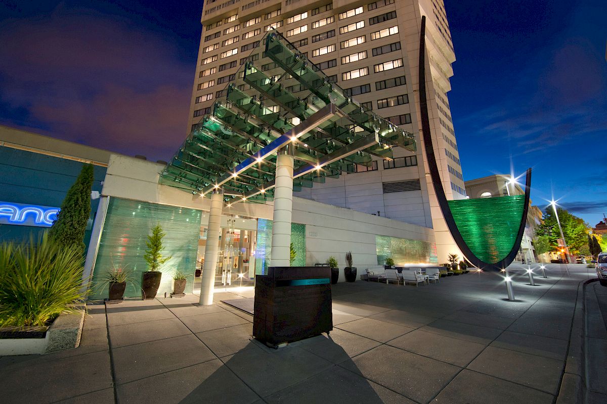 Modern building entrance with glass canopy, decorative green structure, and potted plants on paved area at dusk with lights illuminating the scene.