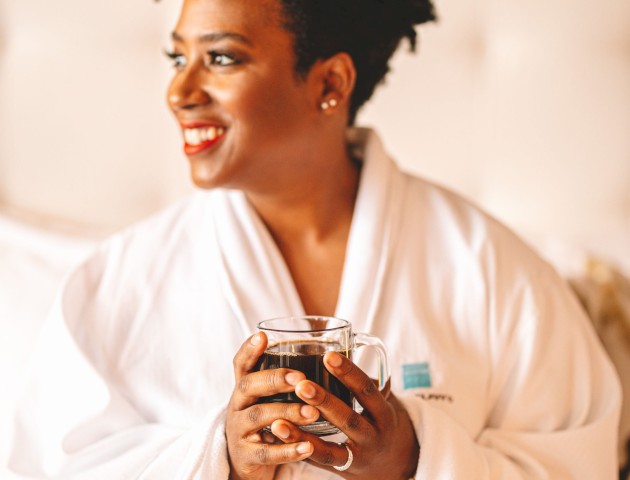 A person wearing a white robe is smiling and holding a cup while sitting on a bed. The background appears to be a bedroom, with a headboard visible.