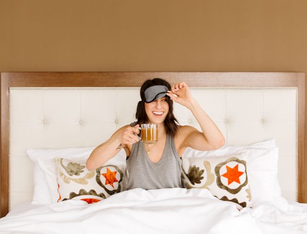 A person in a bed is lifting a sleep mask while holding a glass mug, smiling and sitting under a white blanket with decorative pillows.