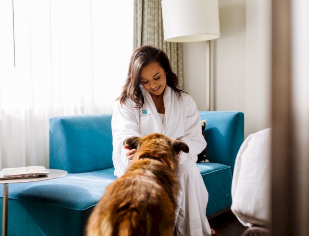 A woman in a white robe is sitting on a blue sofa, smiling and petting a brown dog in a well-lit room.