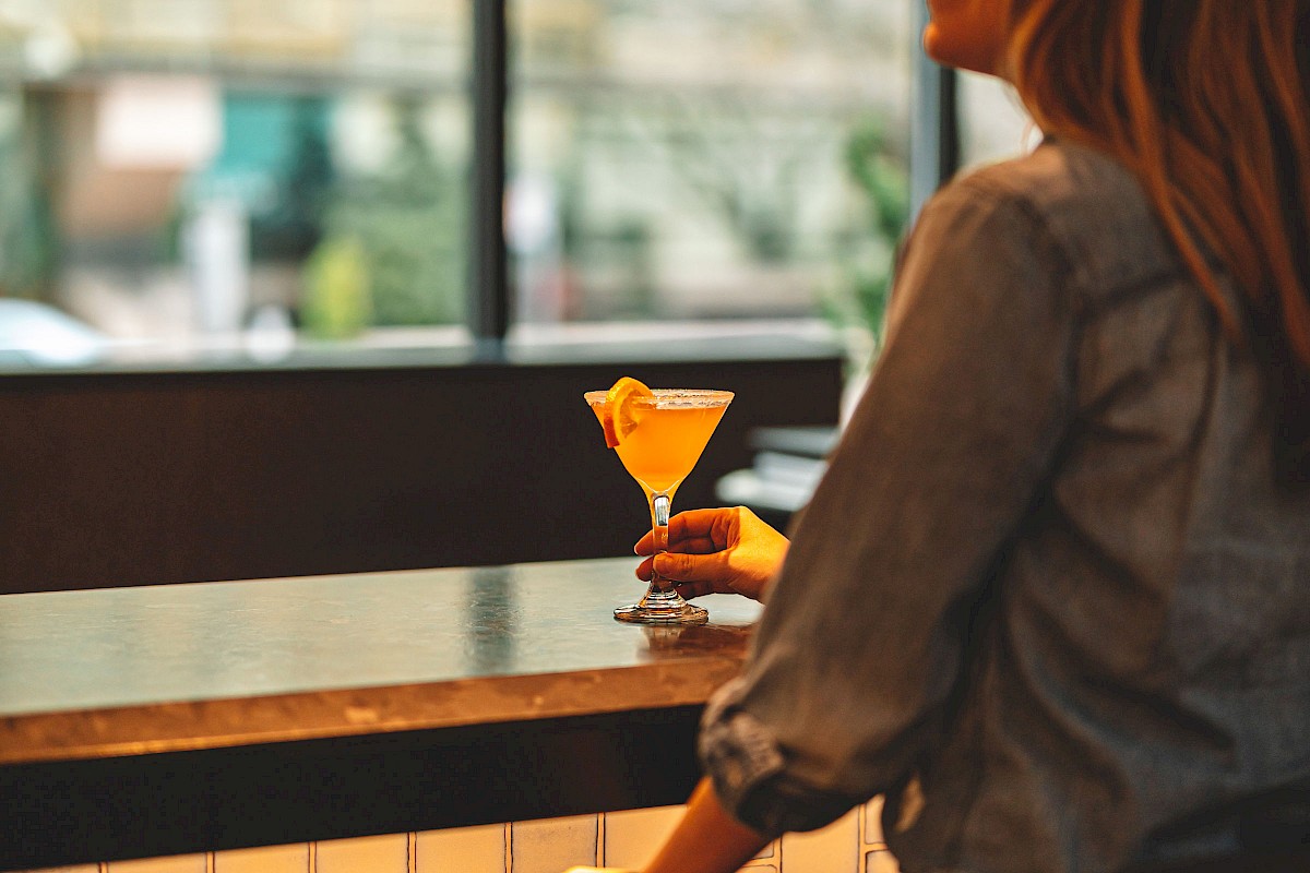 A person sitting at a bar counter, holding a cocktail glass, and smiling in a warmly lit environment with large windows behind.