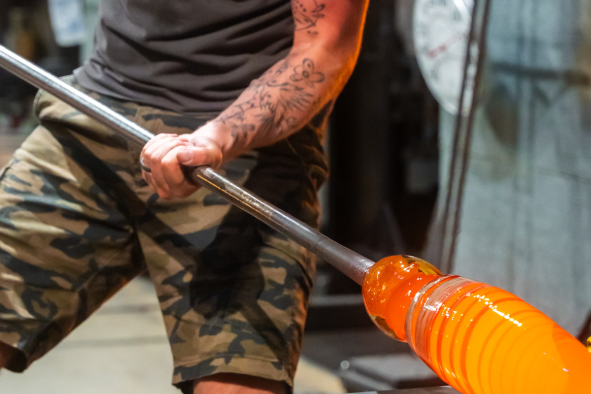 Two individuals are working on glassblowing, shaping molten glass on a rod while wearing protective gear and casual clothing.