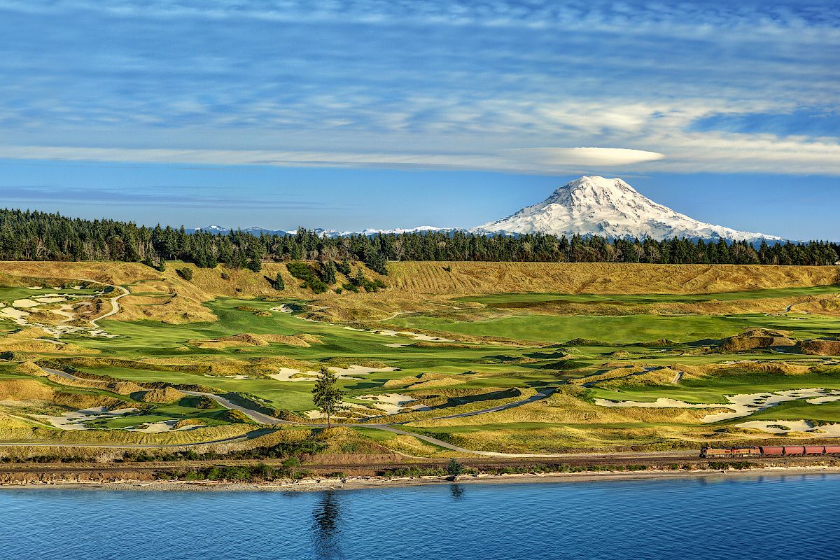 A scenic view of a golf course with a snow-capped mountain in the background, under a partly cloudy sky, with a body of water in the foreground.