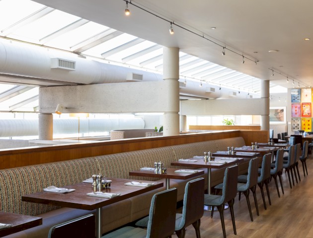 This is an image of a modern, well-lit restaurant interior with tables, chairs, colorful wall art, and a large window skylight ceiling.
