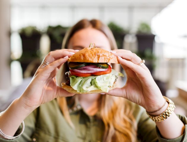 A person holding a large sandwich with various vegetables, including lettuce, tomato, cucumber, and onion, in a blurred background setting.