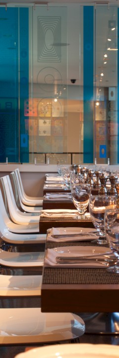 A stylish, modern restaurant interior with a row of white chairs, tables set with glasses, napkins, and salt/pepper shakers, near a colorful glass wall.