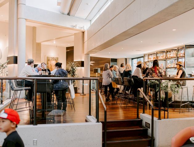 People are gathered in a modern café or bar, engaging in conversation and enjoying drinks. A child in a red cap is also visible in the foreground.