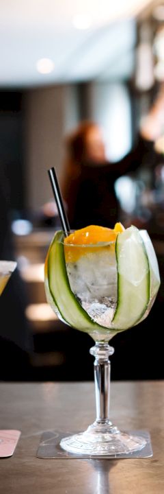 Five cocktails with various garnishes sit on a bar counter with people and a stocked bar in the background.