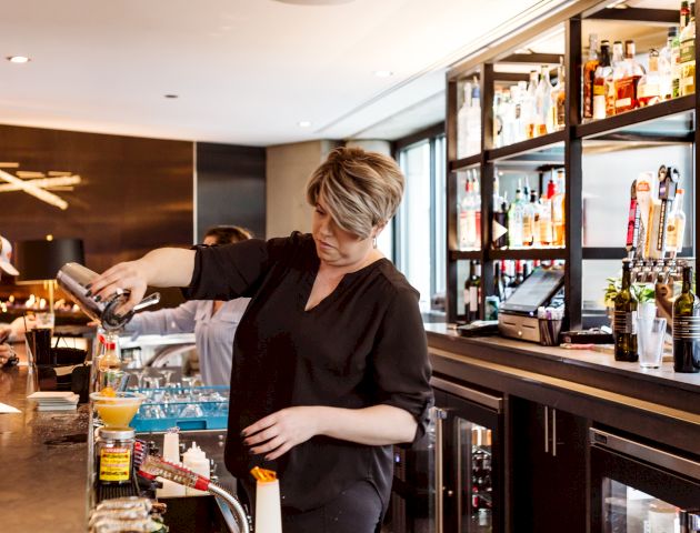 A bartender is pouring a drink from a shaker at a bar, with a variety of bottles and beverages visible on shelves behind her.