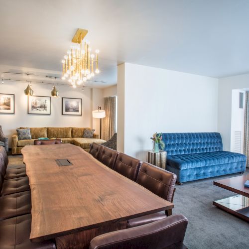 The image shows a modern living room with a long wooden table, blue couch, brown chairs, and wall art, illuminated by a chandelier.