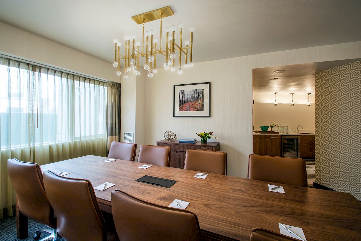 A modern conference room with a long wooden table, brown chairs, a chandelier, and window curtains, with a small kitchenette area visible in the back.