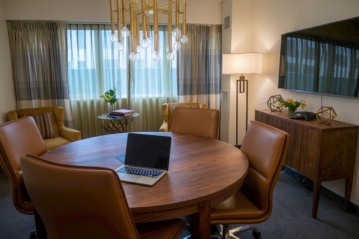A modern, well-lit office space with a round wooden table, leather chairs, a laptop, a chandelier, a TV, and a side table with decor, ending the sentence.