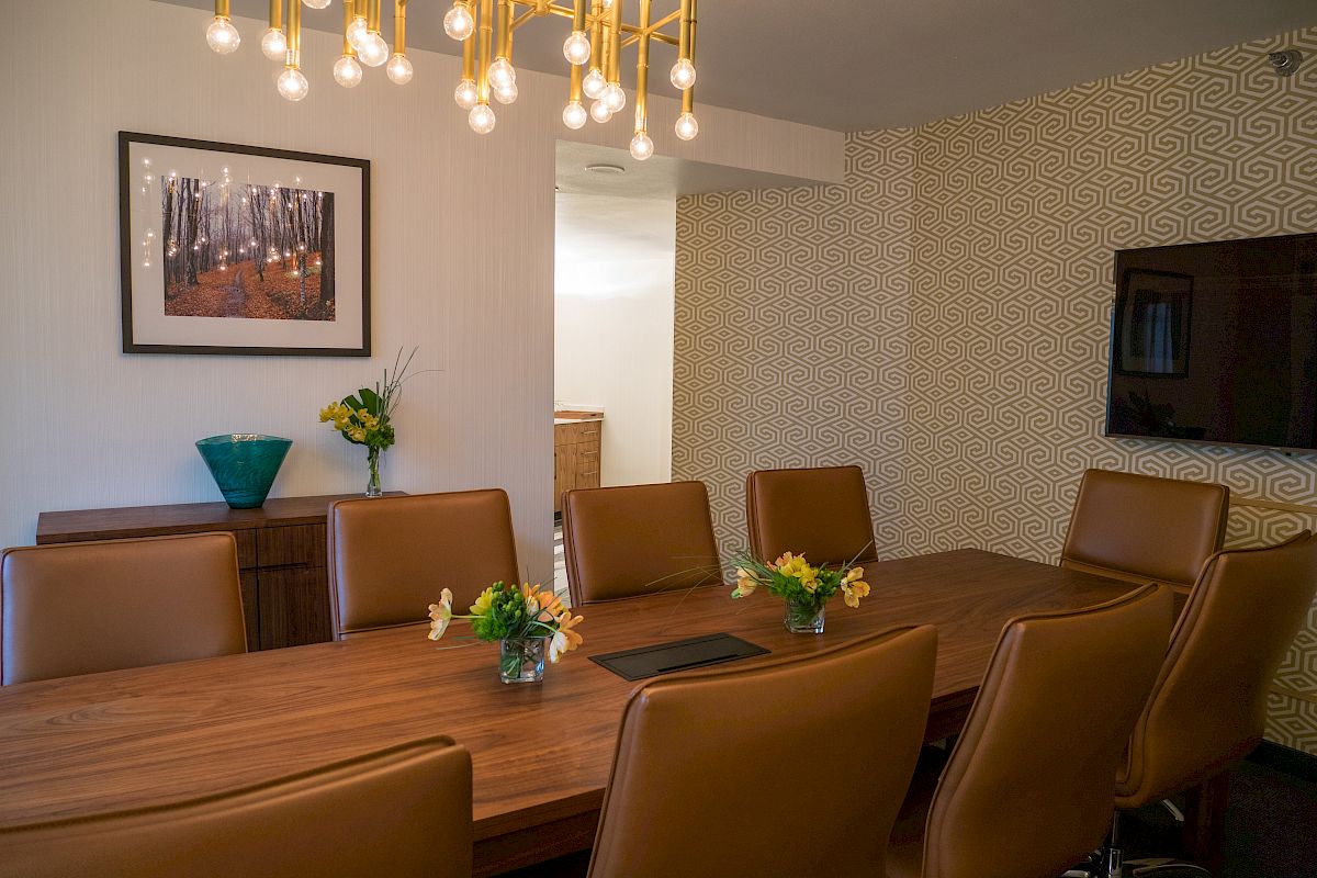 A modern dining room with a wooden table, brown chairs, a light fixture, framed wall art, and floral arrangements, with accent wallpaper and a TV.