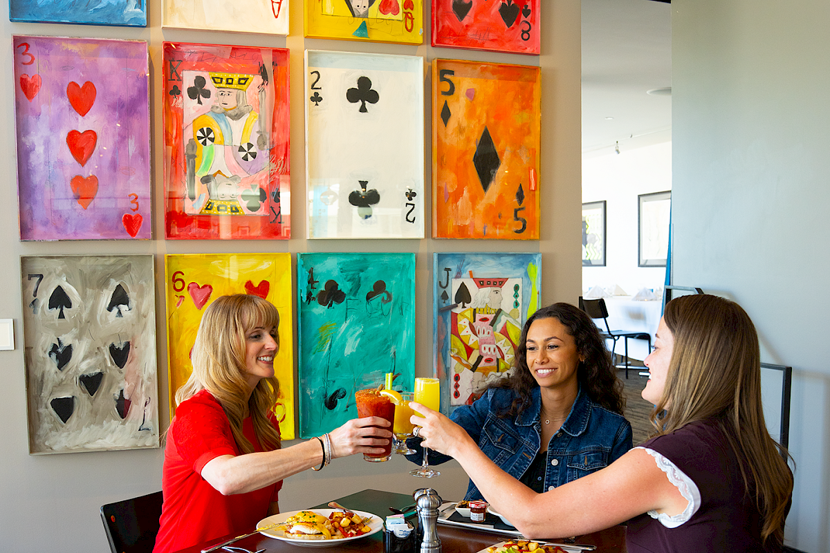 Three women clink drinks at a table in a bright dining space with colorful card-inspired wall art, shown as part of Hotel Murano’s Black Friday 2025 and Cyber Monday cyber sale visuals.