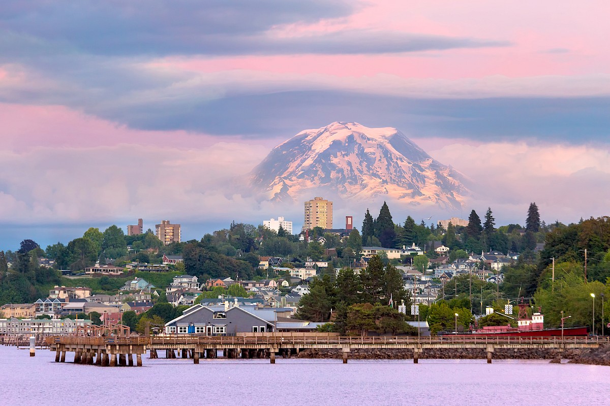 A scenic view of a town with a snow-capped mountain in the background during sunset, featuring a pier, houses, and lush greenery.