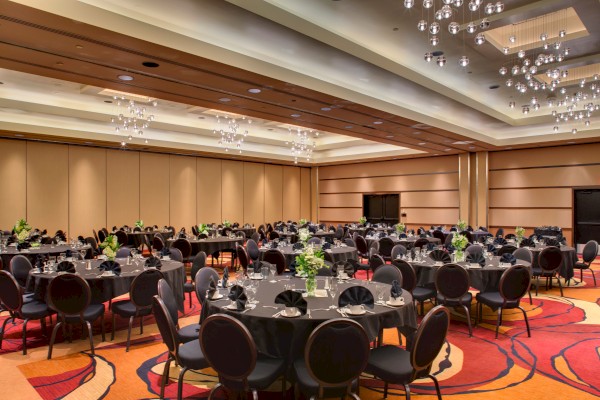 The image shows an elegantly decorated banquet hall with round tables, black tablecloths, chairs, and bright chandeliers overhead.