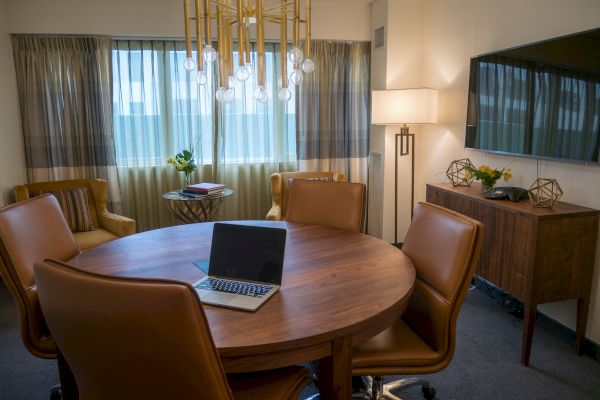 A conference room with a round wooden table, brown chairs, a laptop, a chandelier, a TV, a side table with decor, and a standing lamp.