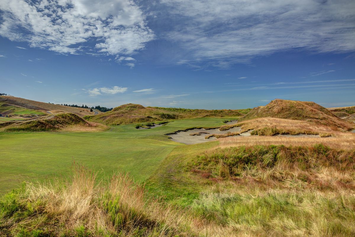 A scenic golf course with grassy hills, sand traps, and a clear blue sky dotted with clouds.