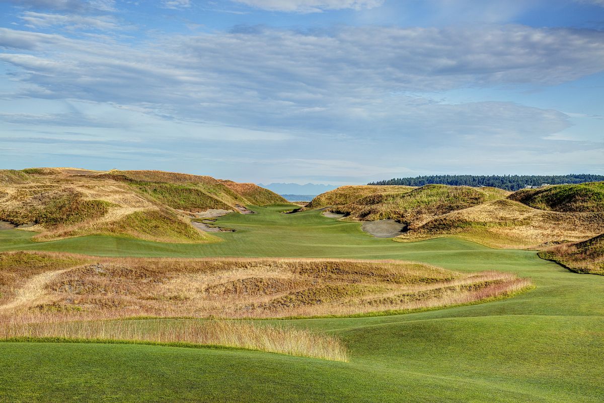 The image shows a scenic landscape of a golf course with green fairways, sand bunkers, and grassy hills under a partly cloudy sky.