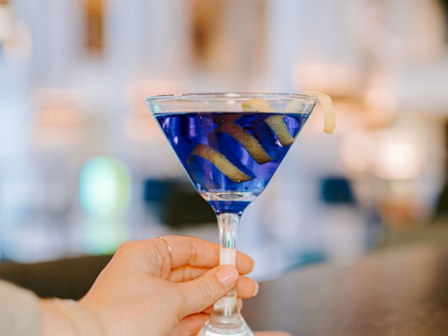 A hand holding a blue cocktail in a martini glass with a lemon twist garnish at a bar, placed on a coaster.