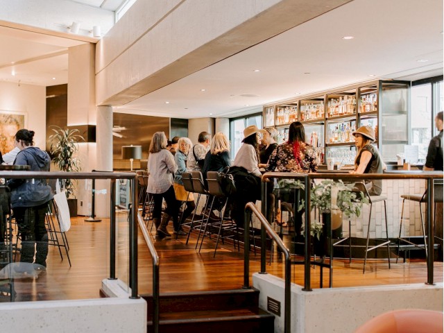 The image shows a modern indoor cafe or bar with several people seated at the counter and tables, engaged in conversation.