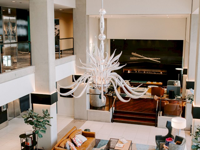 This is an image of a modern hotel lobby with a large chandelier, seating areas, plants, and a fireplace, viewed from an upper floor level.