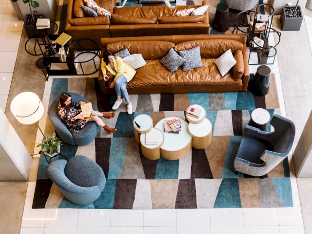A modern lounge area with two people sitting on a leather sofa and a blue chair, surrounded by round tables and stylish decor, on a patterned rug.
