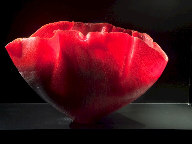 This image shows a vibrant red, artistically crafted bowl on a dark background, illuminated dramatically to highlight its texture and form.