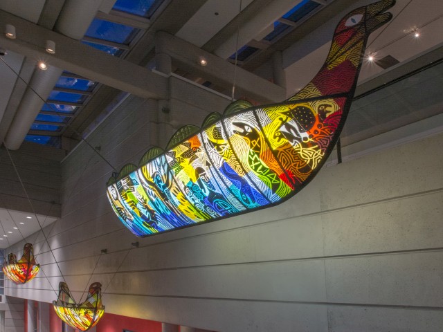 The image shows colorful, intricately designed boat sculptures suspended from a high ceiling in an indoor space, possibly an art installation.