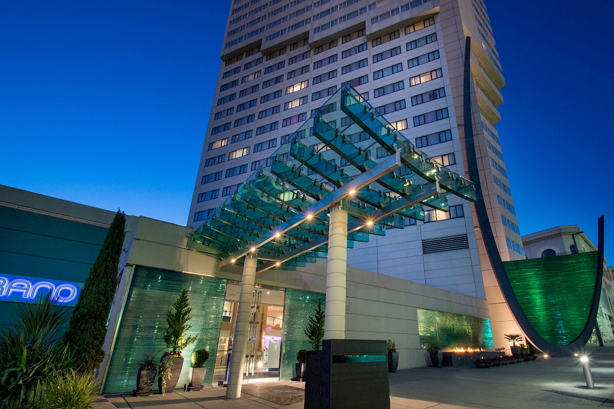 The image shows the entrance of a modern high-rise building with glass and metal architectural elements, illuminated at dusk.