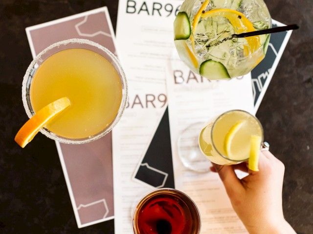 The image shows an overhead view of four cocktails, a hand holding one, placed on menus labeled 