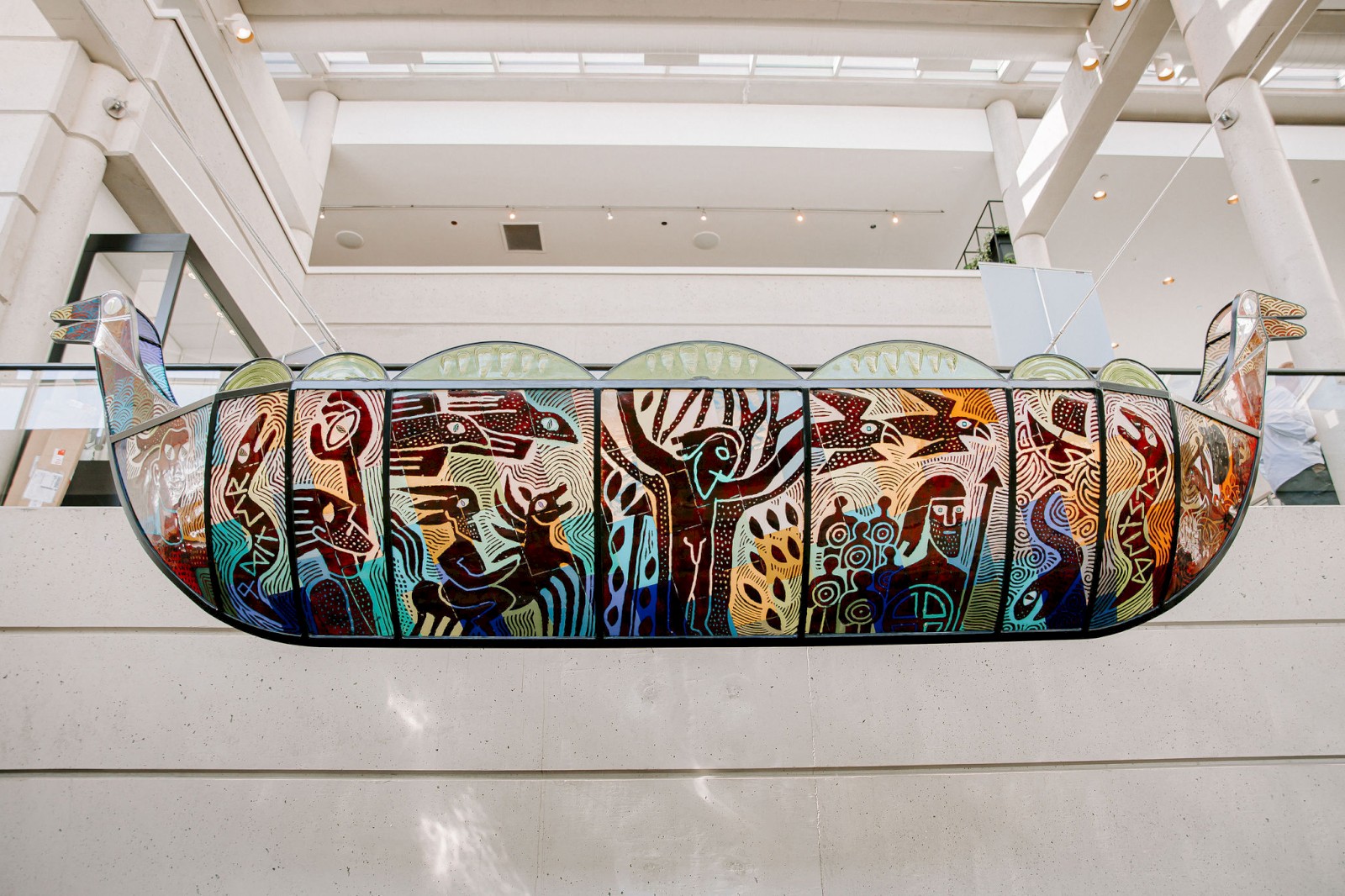 The image shows a stained glass art piece shaped like a boat, with various colorful, abstract designs and figures, hanging in a modern building.