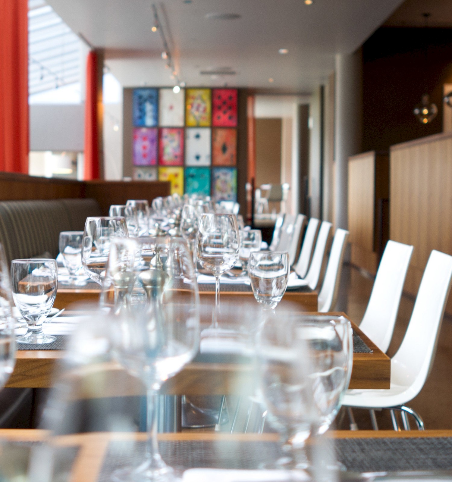 An elegant restaurant with neatly set tables, glassware, and white chairs, featuring modern decor and colorful art in the background.