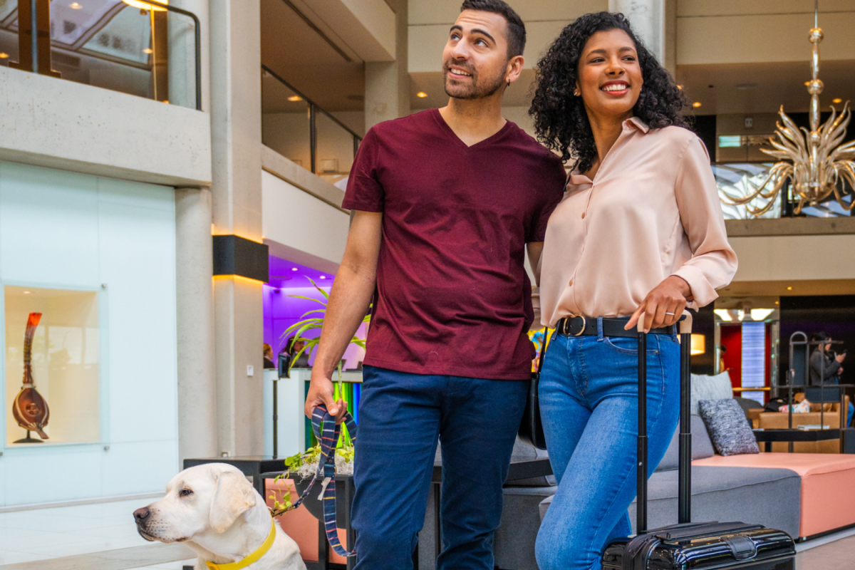 A man and woman stand in an open, modern interior. The man holds a guide dog, and the woman has her hand on a suitcase handle.