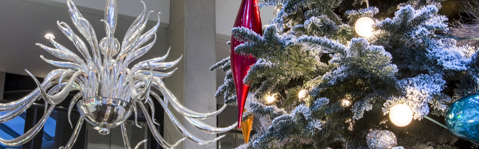 A decorated Christmas tree adorned with lights and ornaments stands next to an intricate chandelier in an indoor setting.