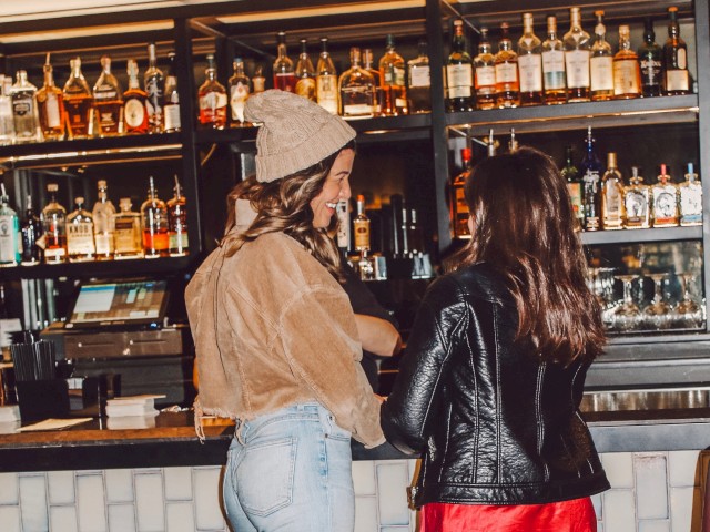 Two people standing at a bar with various bottles on shelves behind them.
