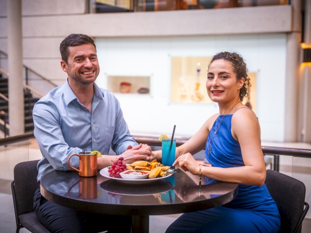 Two people are sitting at a table with drinks and snacks, smiling at the camera in a modern indoor setting.