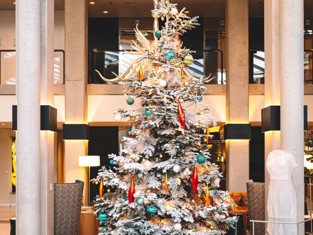 A decorated Christmas tree with lights and ornaments stands tall in a modern indoor setting, flanked by columns and plush chairs.