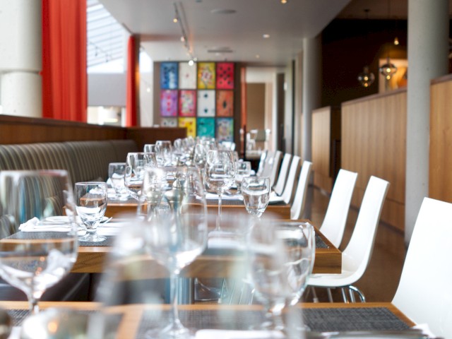 A modern restaurant with neatly arranged tables, stemware, and colorful wall art. White chairs and wooden decor create a stylish ambiance.