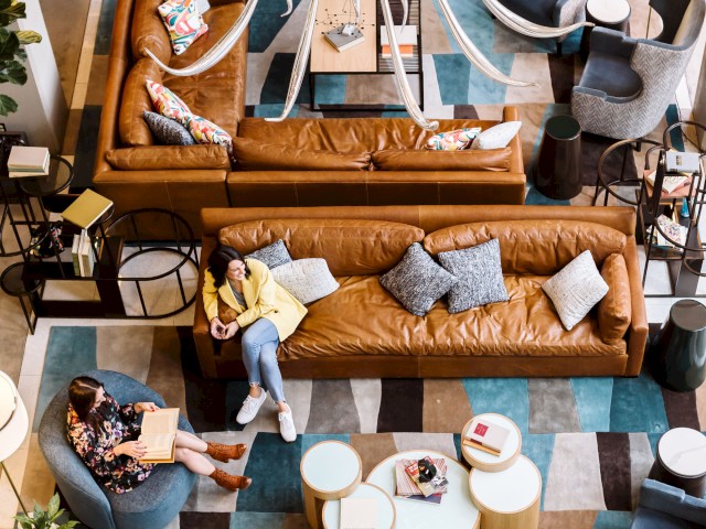 A stylish lounge with brown sofas, blue-gray chairs, a colorful rug, abstract chandelier, and a few seated people reading or relaxing.